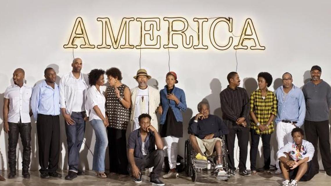 Under Glenn Ligon’s “America” a group of 30 Americans artists, left to right: Rashid Johnson, Nick Cave, Kalup Linzy, Jeff Sonhouse, Lorna Simpson, Carrie Mae Weems, Barkley L. Hendricks, Hank Willis Thomas (front row), Xaviera Simmons, Purvis Young, John Bankston, Nina Chanel Abney, Henry Taylor, Mickalene Thomas (front row), Kerry James Marshall, and Shinique Smith.
