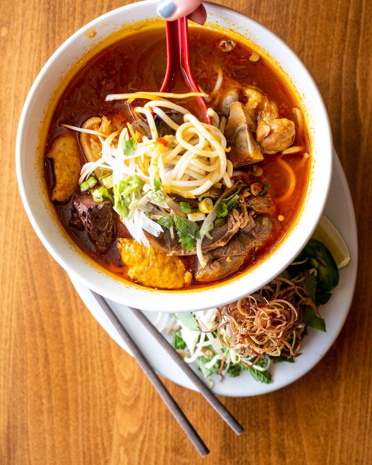 Deep-red, compliments of chilis and congealed blood, a beef and pork broth joins shrimp paste, pig’s feet, beef shank and homemade pork sausage to make Lunar Pho’s bún bò Huế. The plate includes banana flowers, expensive but essential to the dish.
