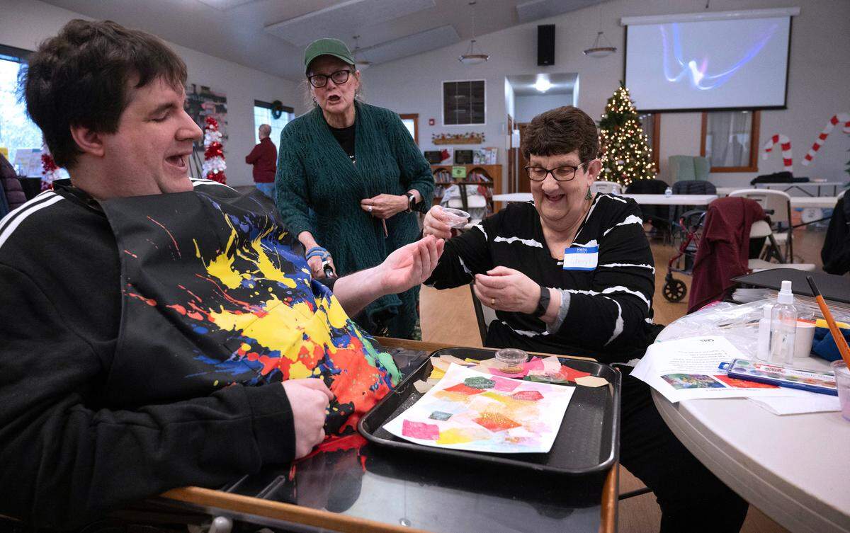 With the assistance of his caregiver and mother Cheryl Cleveland, Nathan Tripp of Edgewood sprinkles glitter over his watercolor and paper artwork during the Lutheran Community Services’ Opening Minds Through Art program at the Mountain View Community Center in Edgewood, Washington, on Wednesday, Dec. 6, 2023.