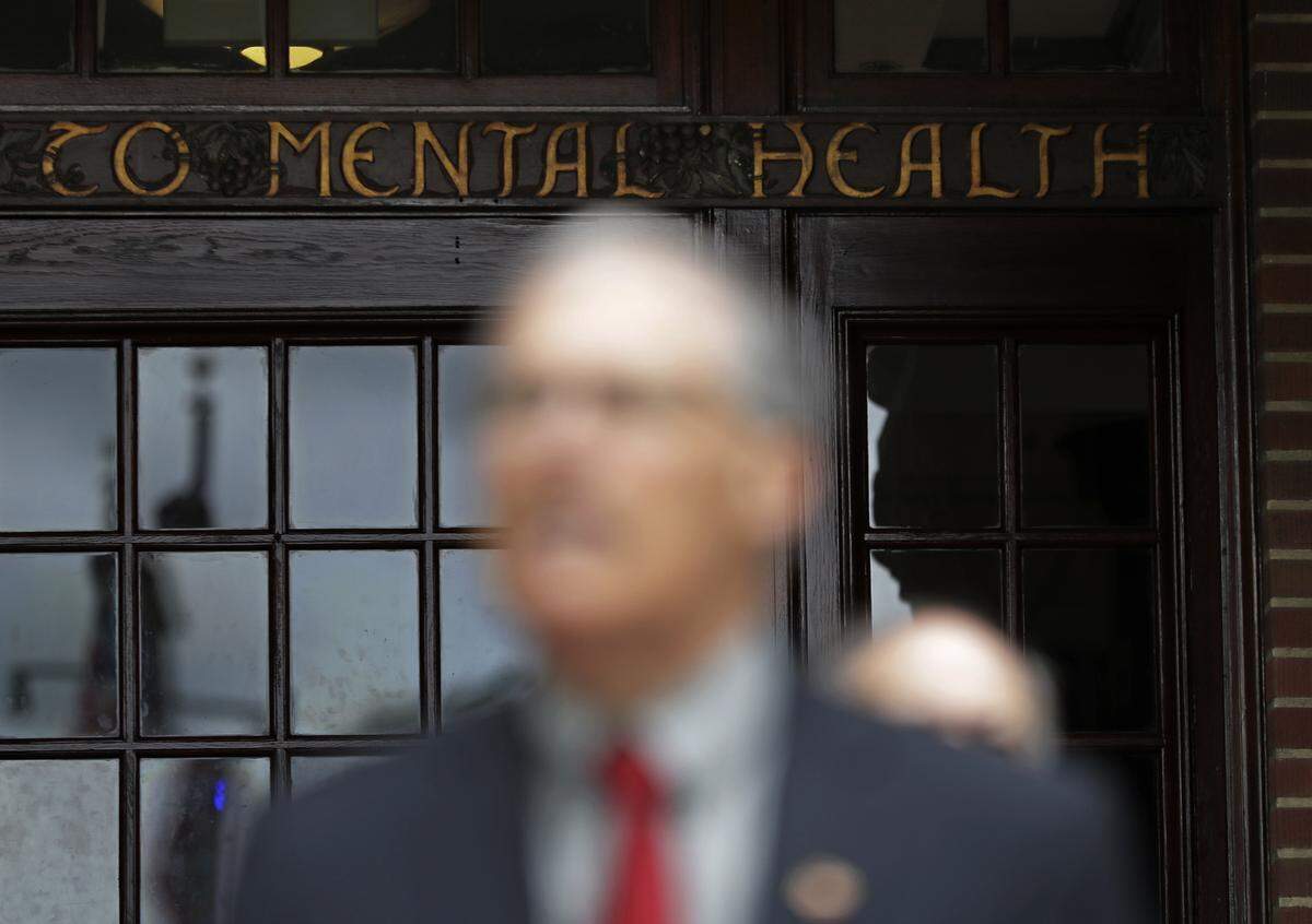 A portion of a wooden sign that reads "Dedicated to Mental Health" above an entrance to Western State Hospital in Lakewood is shown as Washington Gov. Jay Inslee speaks Friday, May 11, 2018 during a news conference.