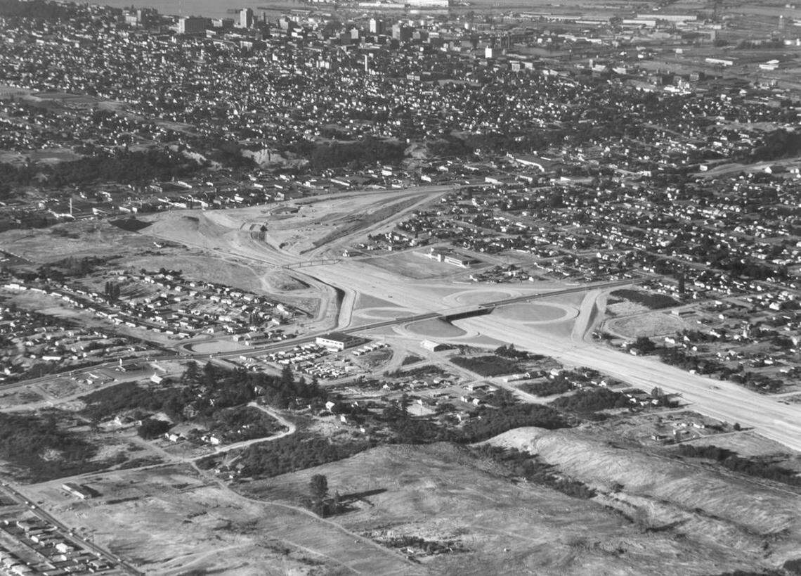 This aerial photograph taken Aug. 17, 1960 shows the I-5 interchange at South 38th Street. The excavation work has begun, and the 38th Street bridge over I-5 is complete. The building to the left of the cloverleaf is Langendorf Bakery, 2202 S. 38th St., which was built in 1954. The large area of cleared land at the bottom of the photograph is the future home of the Tacoma Mall. Part of downtown Tacoma and the tideflats can be seen at the top edge of the picture.