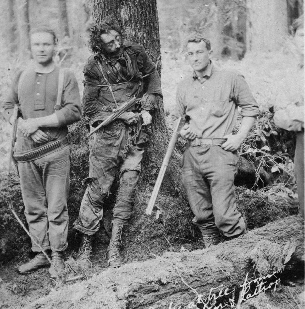 A postcard captioned, “Tornow’s Body at tree from which he shot Blair and Lathrop”. Two men with rifles pose, one on each side of Tornow’s corpse, in whose dead arms they have propped a rifle. The man standing on the left is wearing an ammunition belt around his waist.