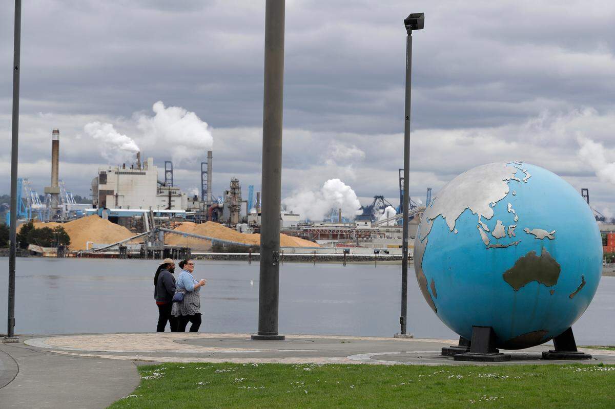 In this April 21, 2020 photo, people walk past an Earth globe sculpture at Thea’s Park in Tacoma, Wash., with the WestRock Paper Mill in the background. (AP Photo/Ted S. Warren)