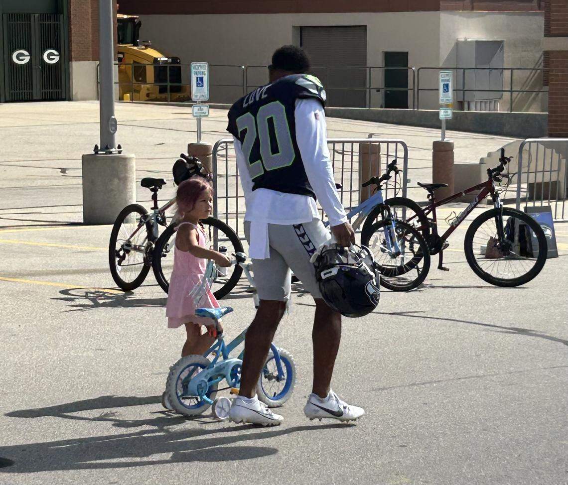 Seahawks safety Julian Love (20), a father of a young child, thrills a local girl by choosing her small bike over bigger bikes from bigger kids to ride from the locker room at Lambeau Field to the practice fields for Seattle’s joint practice with the Green Bay Packers in Wisconsin Thursday, Aug. 21, 2025.