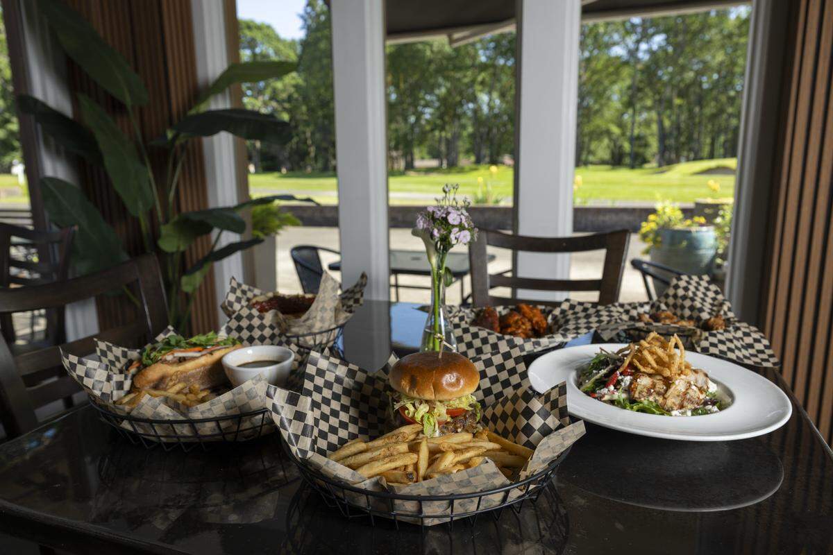 A table filled with selections from The Edit Social House's menu, including a Banh Mi Dip, left, True Smash Burger, center, and a seasonal salad, right, overlooks the Oakbrook Golf Club green on Thursday, May 21, 2026, in Lakewood, Wash.