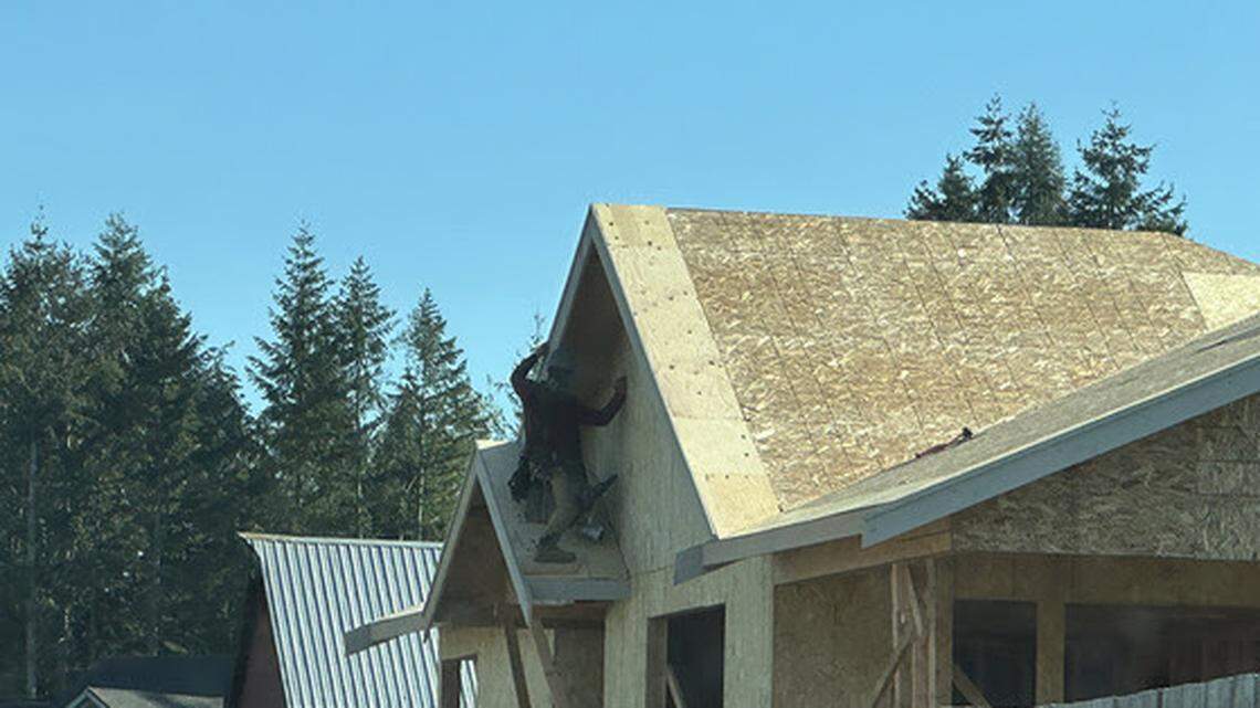 A Modern S Construction worker who is not wearing fall protection is standing on a small roof awning, holding onto the main roof with one arm.