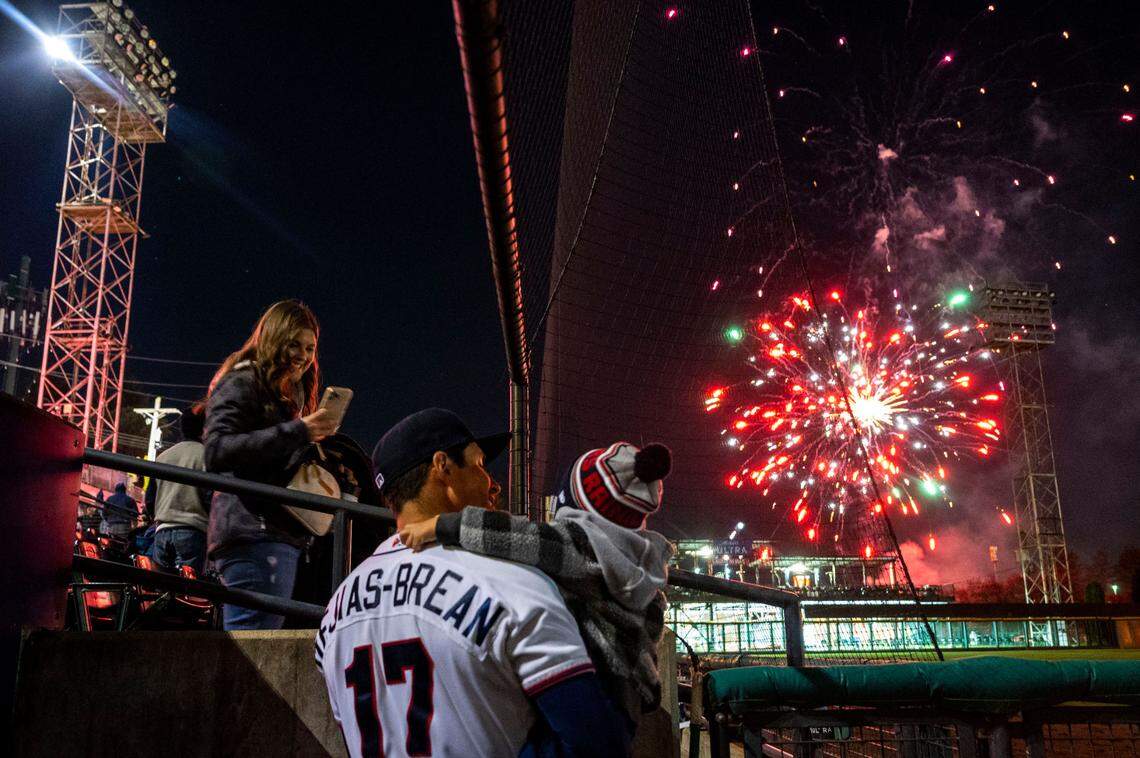 Tacoma first-base coach Seth Mejias-Brean (17) holds his son, Beckett, as his wife, Haley, looks on during the post-game fireworks show after the season opener at Cheney Stadium in Tacoma, Wash., Tuesday, April 5, 2022. Tacoma beat the Salt Lake Bees, 4-2.