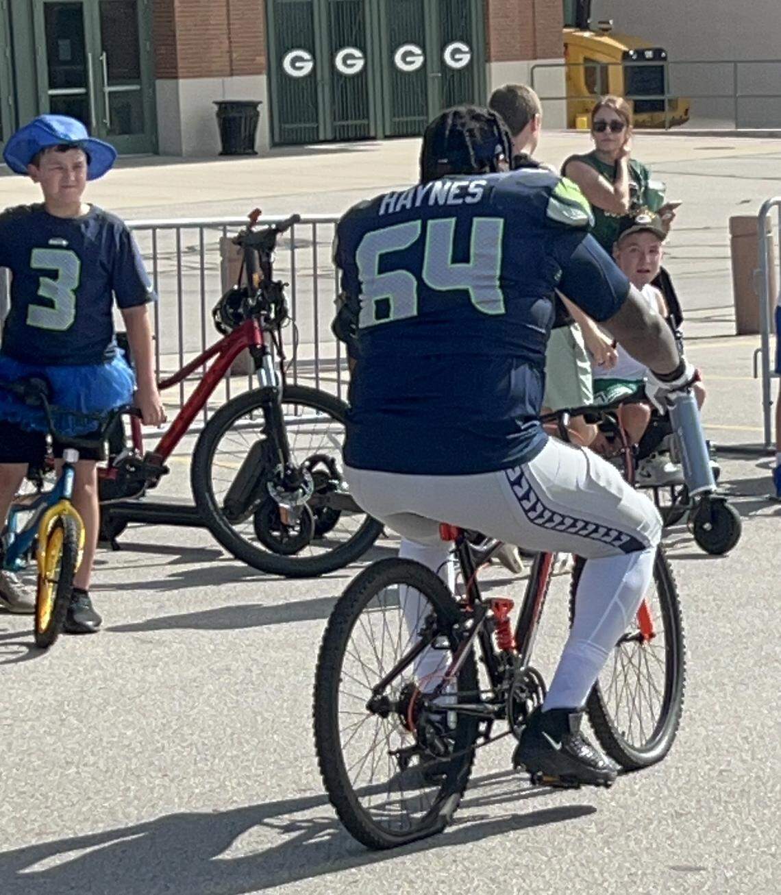 Seahawks 317-pound guard Christian Haynes flattens the back tire of a Green Bay boy’s bike he borrowed to ride to Seattle’s NFL preseason joint practice with the Packers outside Lambeau Field in Green Bay, Wisconsin, Thursday, Aug. 21, 2025.