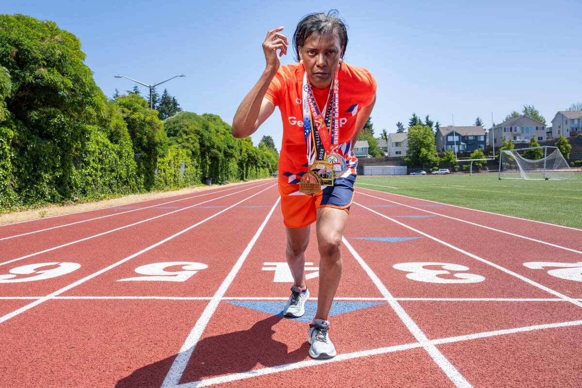 Madonna Hanna poses for a portrait in a sprinting stance, wearing medals from competing in the National Senior Games, on Tuesday, July 8, 2025, on the track at Truman Middle School in Tacoma.