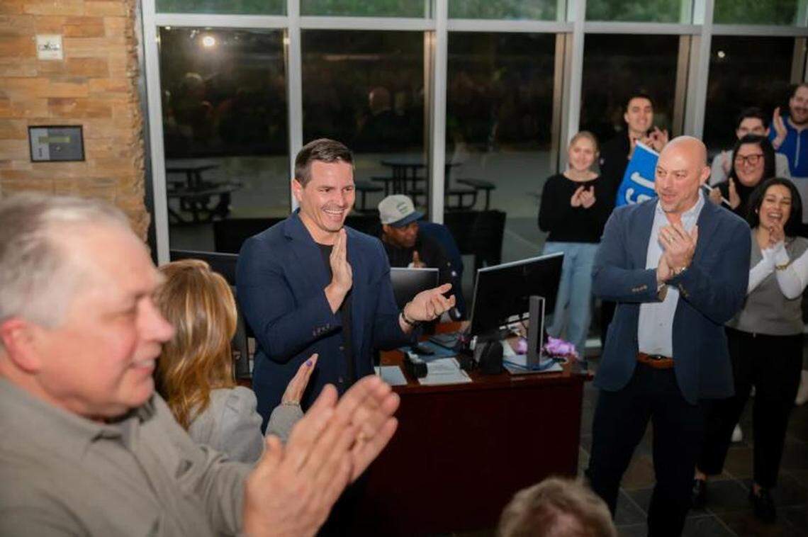 With team vice chair Bert Kolde (foreground left) and team president Chuck Arnold (right, hands clapped, dark-blue jacket over light shirt) flanking him, new Seahawks coach Mike Macdonald (center) gets a rousing welcome within his first steps inside the franchise’s Virginia Mason Athletic Center Wednesday, Jan. 31, 2024.