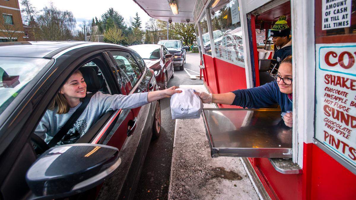 Bella Hinton, right, hands Shawna Cole her order as cars queue up at the Frisko Freeze drive-thru window on April 8, 2020, before a statewide mask mandate went into effect. The 70-year-old fast food icon is weathering the coronavirus crisis the same way it always has – serving up Tacoma’s favorite greasy burgers and fries.