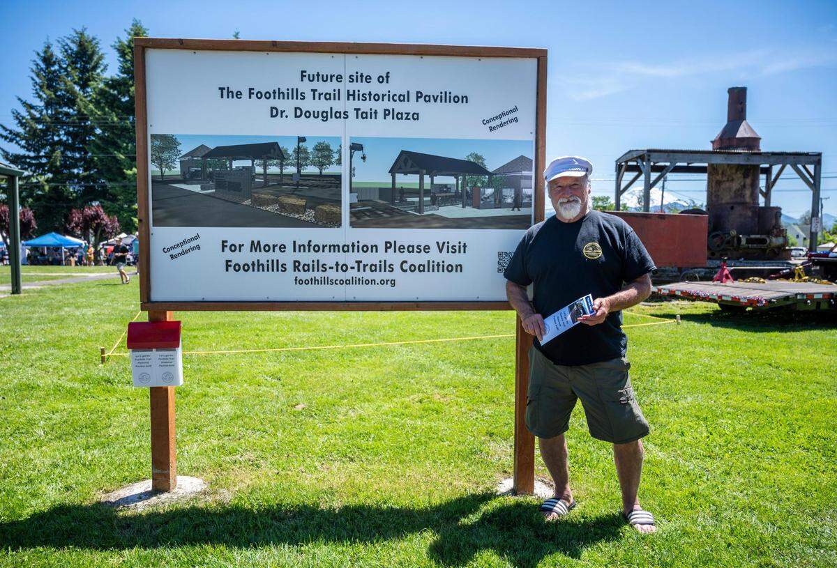 The Foothills Rails-to-Trails Coalition committee member Milt Tremblay stands next to a sign that displays what a future pavilion in honor of Dr. Douglas Tait will look like once it’s built behind Wally’s Drive-In in Buckley, Wash. on Saturday, June 25, 2022.