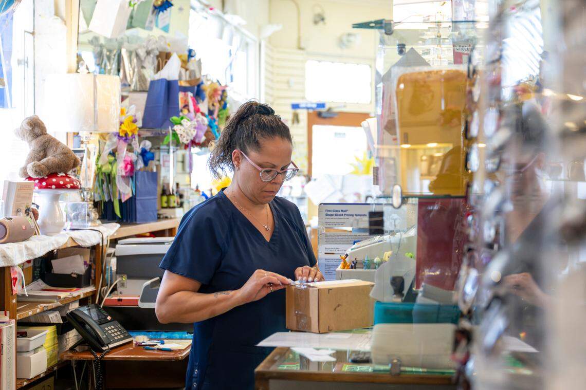 Alicia Mendez places an address sticker on a package from behind the counter of the USPS substation at Rankos’ Pharmacy on Wednesday, July 9, 2025, in the Stadium District of Tacoma. Mendez works as a pharmacy assistant, while operating the post office at the pharmacy.