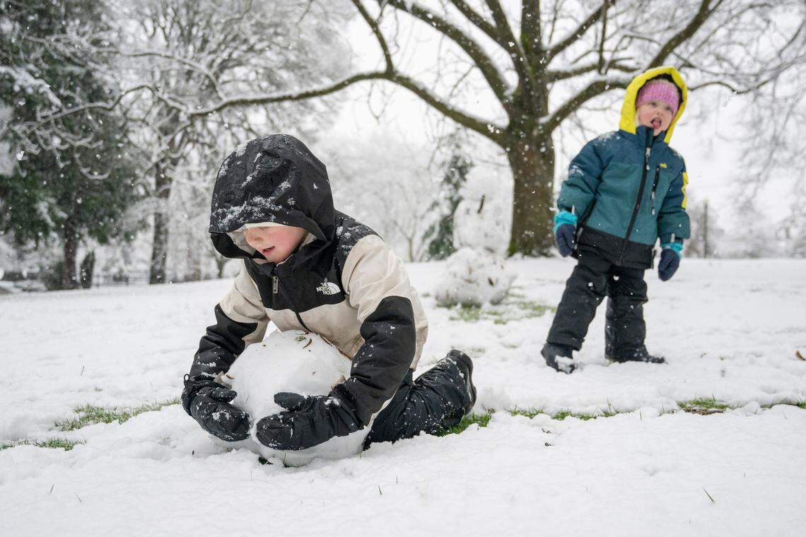 Eli, 7, rolls the base of a “snow baby” to join an already built snowman as Lucy, 3, sticks a tongue out to taste falling snow on Friday, March 13, 2026, at Wright Park in Tacoma, Wash.