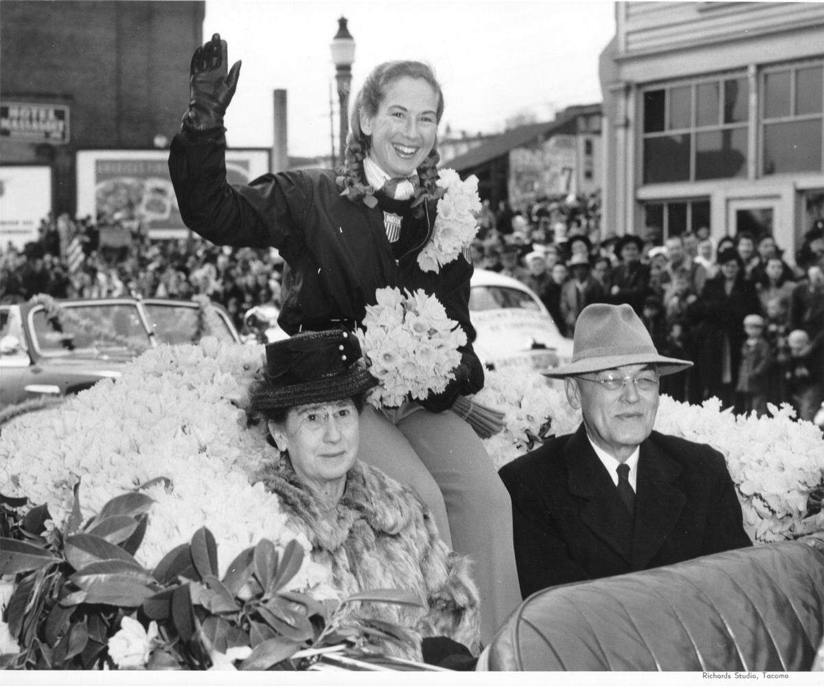 Gretchen Kunigk Fraser, winner of Gold and Silver medals at the 1948 Winter Olympics, waves to thousands of spectators viewing the April, 1948 Daffodil Parade. She is wearing her U.S. team uniform and her familiar blond pigtails. Gretchen was the first American skier to medal in a Winter Olympic event with a win in slalom and a silver in the Alpine combined at St. Mortiz. Her parents, W.A. and Clara Kunigk, ride along surrounded by mounds of daffodils. (Tacoma Public Library)