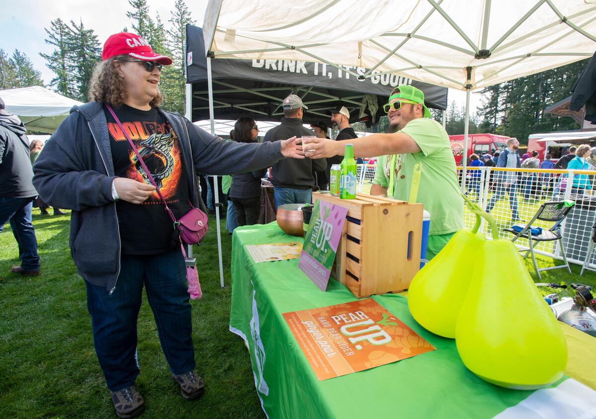 Pear Up owner Kevin Van Reenen of Wenatchee hands a sample of his ginger pear hard cider to Jeannette James of Tacoma at Cider Swig 2019. The event outgrew its former venue of Sehmel Homestead Park and has moved to LeMay Car Museum in Tacoma for 2022.