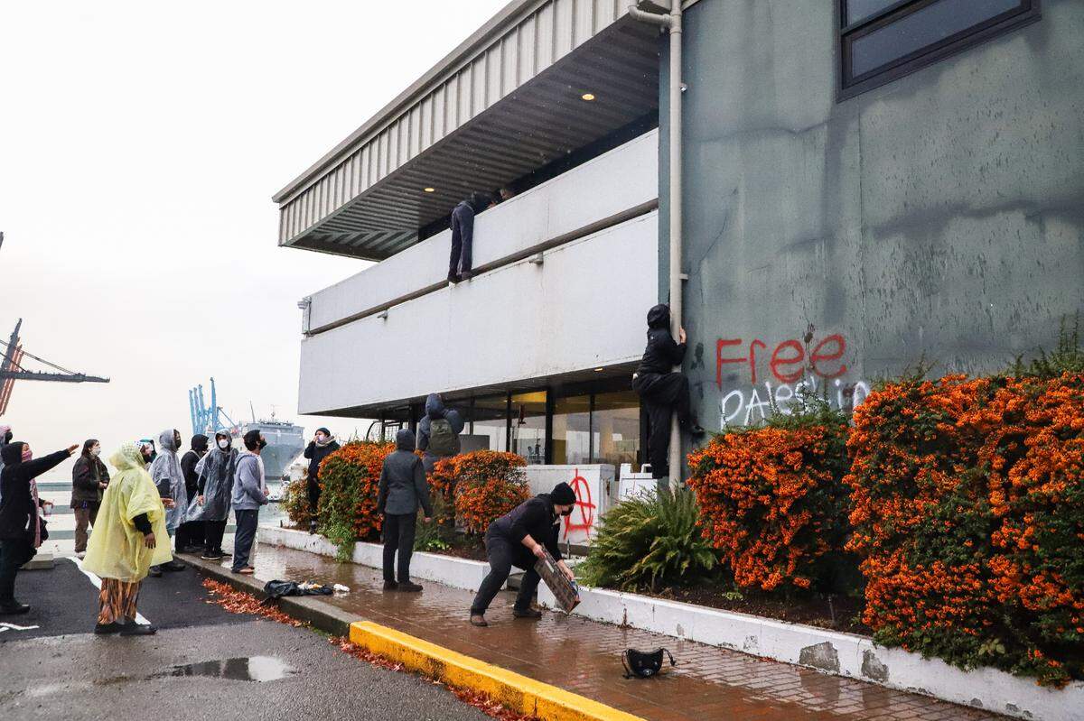 A protester climbs onto the balcony of The Northwest Seaport Alliance building at the Port of Tacoma and a security guard attempts to pull him over the balcony, while a group of protesters scream at the guard to let him go, Monday, November 6, 2023.
