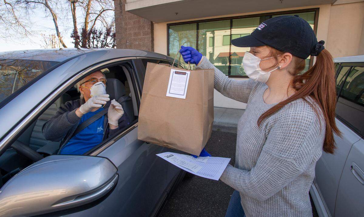 Wearing protective gear, Toscano’s waitress Brianna Saylor brings Craig Knowlton’s order to his vehicle on Tuesday, April 15, 2020. The Puyallup restaurant offers curbside takeout via phone orders to minimize contact between staff and customers. Knowlton, a 78-year-old Edgewood resident, said he and his wife ordered the meal to celebrate their 27th anniversary and was “one of my few adventures outside of the house.”