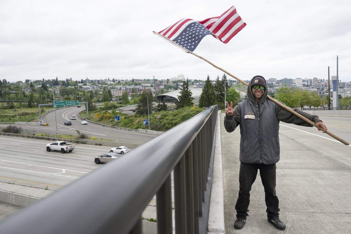 Shaun Anderson poses for a portrait on overpass above Interstate 5 on Wednesday, May 6, 2026, in Tacoma, Wash. Anderson holds out a peace sign and an upside down American flag.
