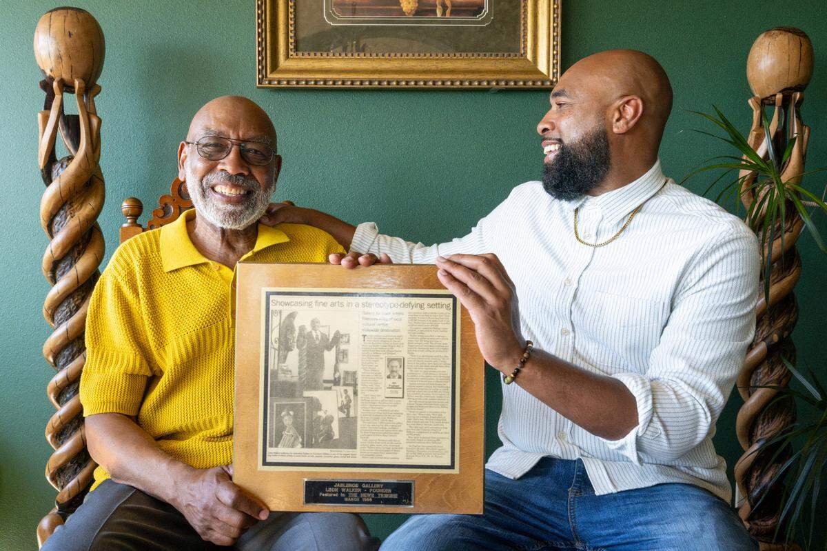 Father and son, Leon Walker, left, and Rob Young, hold a 1996 News Tribune article about JasLeRob Gallery featuring Walker, who founded the African American gallery, as they sit together in Walker’s home on Monday, June 9, 2025, beside the same ebony sculptures by a Benin artist pictured in the original article in Tacoma, Wash. The pair will perform “Said By The Father and Son,” a blend of live hip hop, soul and gospel music and spoken poetry, at the Spanish Ballroom at Tacoma’s McMenamins Elks Temple on June 15.