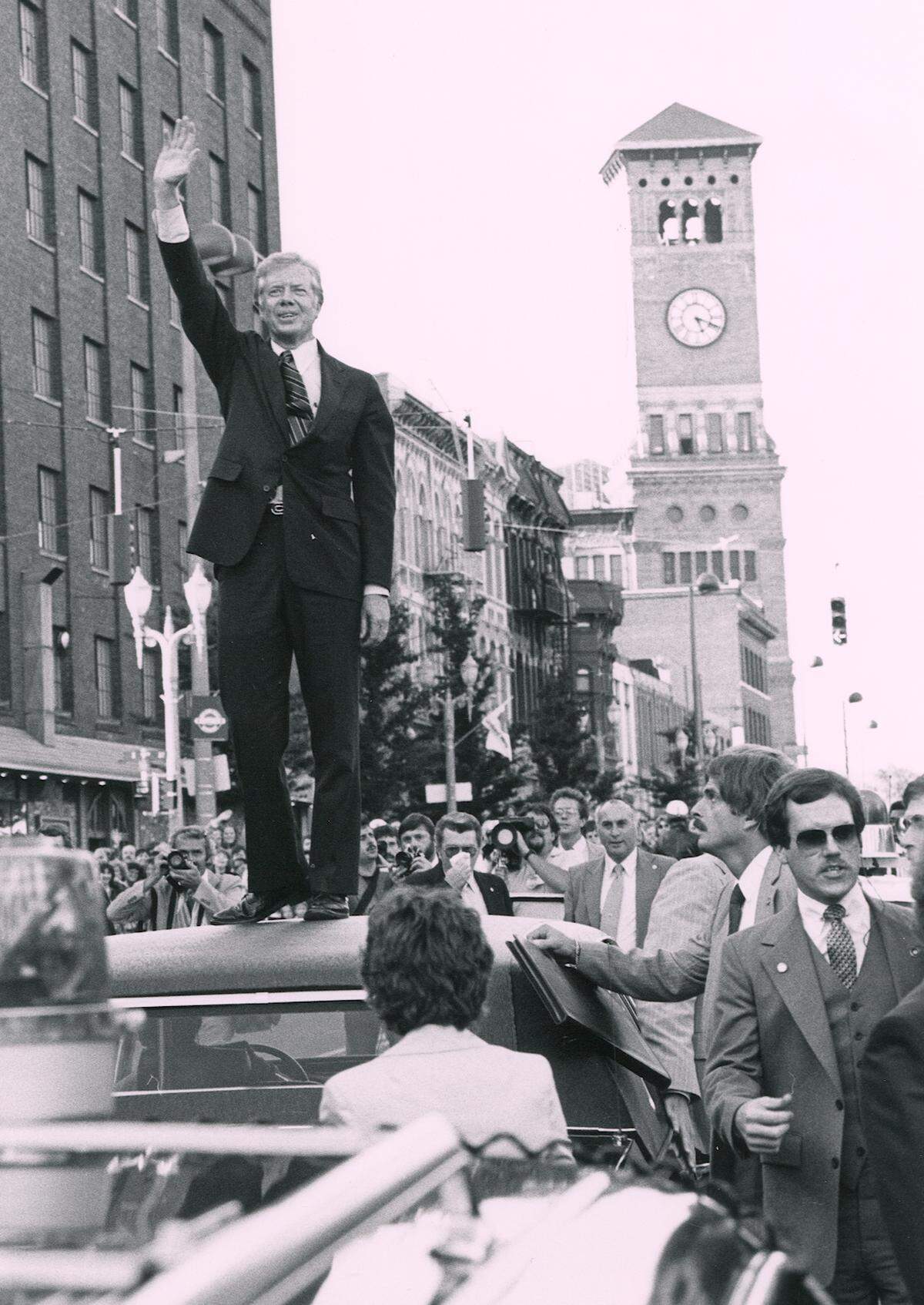 President Jimmy Carter, much to the consternation of his Secret Service agents, has unexpectedly climbed onto the rooftop of his armored limousine as it stopped in downtown Tacoma. He turned and waved to the crowds lining Pacific Avenue for about 30 seconds before descending. Those stuck in the back of the 15-deep crowds were thus able to get a clear view of the President. President Carter then shook as many hands as possible outside the Carter-Mondale campaign office at 9th & Pacific.