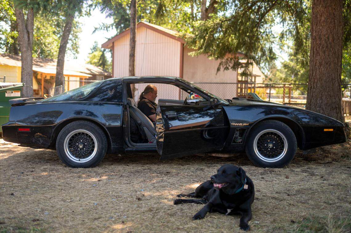 Dennis Casey sits in the drivers seat of his Pontiac Trans AM, while his dog, Diesel, lays in the shade at a safe parking lot at a church in Spanaway, Wash., on Wednesday, July 19, 2023. Casey, and his son, Jerry, have been staying at the safe lot behind the church since April. They have a place to cook food, plug in their phones and a fenced area to live with some piece of mind that their belongings won’t be stolen.
