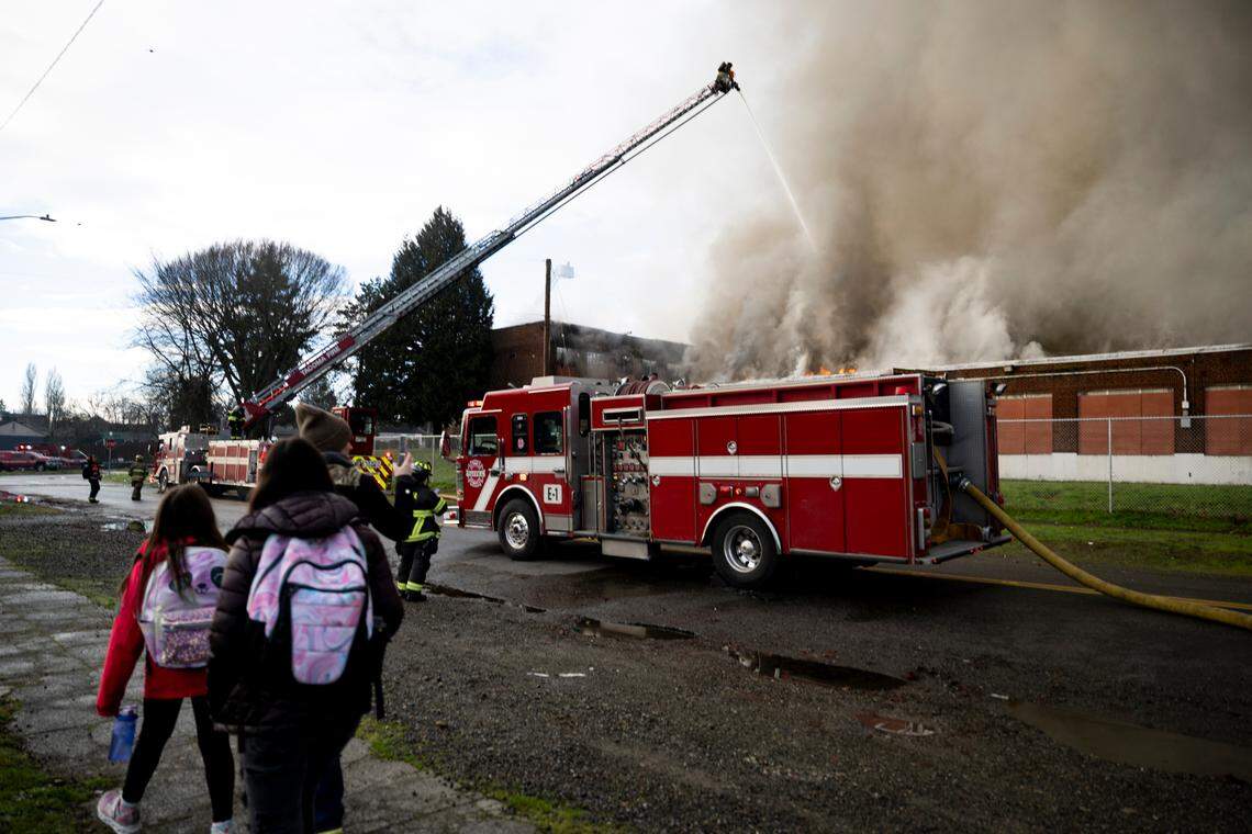 People walk by on their way to school as Tacoma Fire Department Crews battle a fire in the former Gault Middle School building, on Wednesday, Jan. 3, 2024, in Tacoma, Wash. Crews responded around 4:14 a.m. and continue to douse it after 10 a.m.