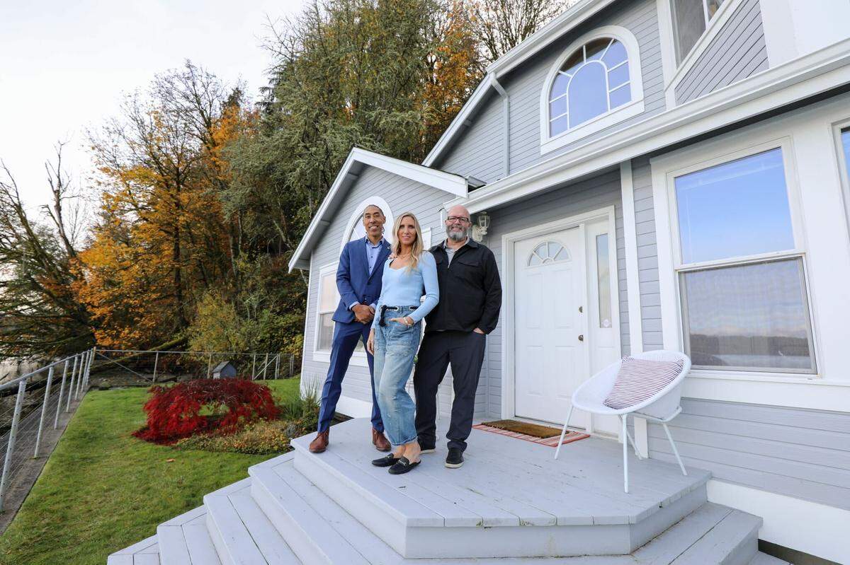 A couple featured on “House Hunters: Amazing Water Homes” pose with their realtor in front of a home facing the waterways of the Puget Sound in Ollala, Washington.