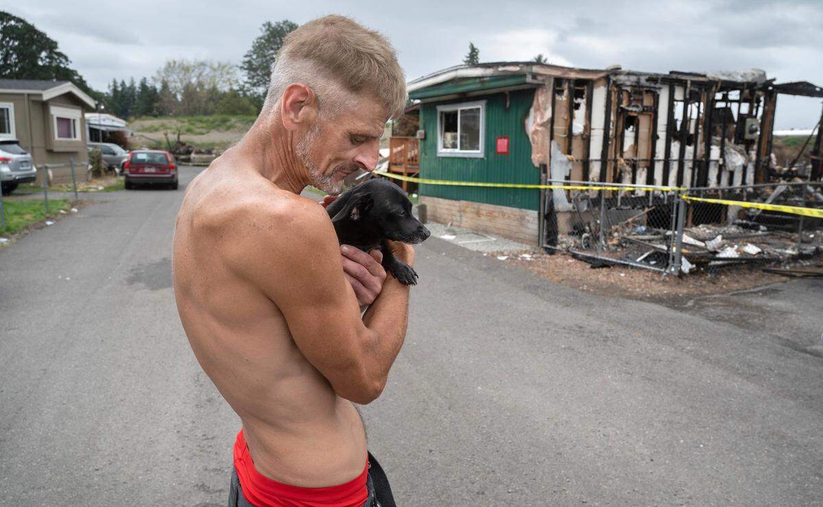 John McLaughlin holds Azee the neighbor’s dog at the Jamestown Estates mobile home park in Lakewood, Washington, on Wednesday, Aug. 9, 2023. McLaughlin used a garden hose to fight off the flames and save his home during last week’s wildfire that killed two men and destroyed nine homes.