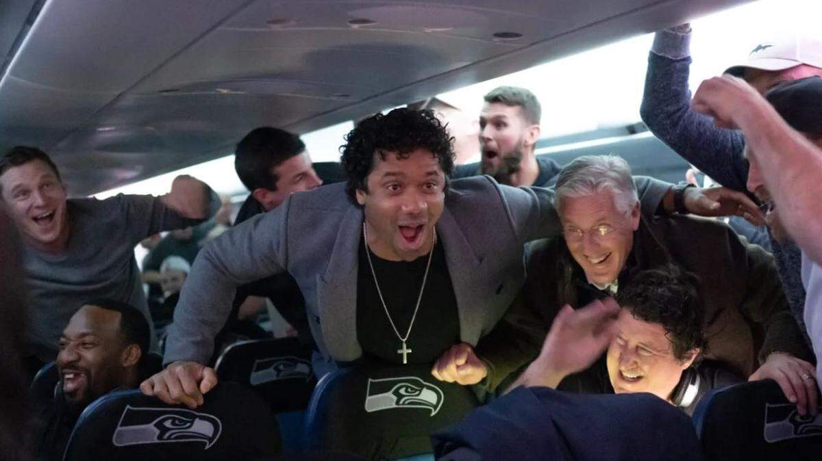 Russell Wilson (center), Pete Carroll (right) and the Seahawks erupt in cheers aboard their chartered jet taking them home from their win at Carolina Sunday, after they learned the Atlanta Falcons beat the San Francisco 49ers. The Niners’ loss put Seattle (11-3) back in first place in the NFC West with two regular-season games remaining.