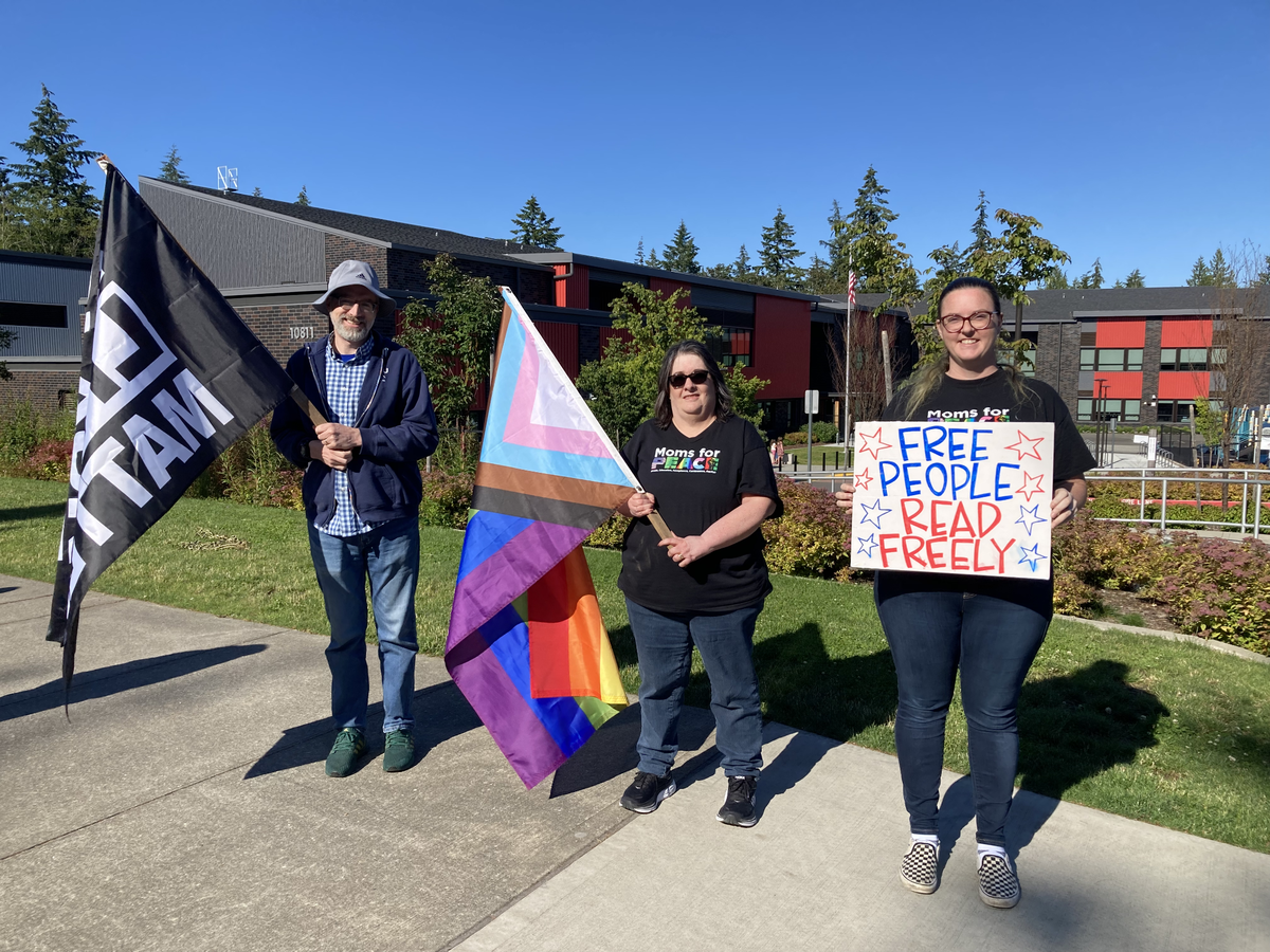 From left to right: Gig Harbor for Racial Justice member Peter Fraser and Moms for P.E.A.C.E. board members Darcy McKnight and Cristy Wahala stand on the sidewalk with flags and signs outside Swift Water Elementary before the Peninsula School District Board meeting on June 18, 2024.