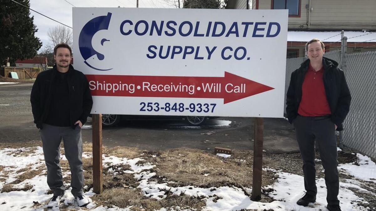 Estimator Travis Martin, left, and Branch Manager Kyle Barrette, right, stand in front of the Consolidated Supply Co. sign in Puyallup. This year, the company is celebrating its 90-year anniversary.