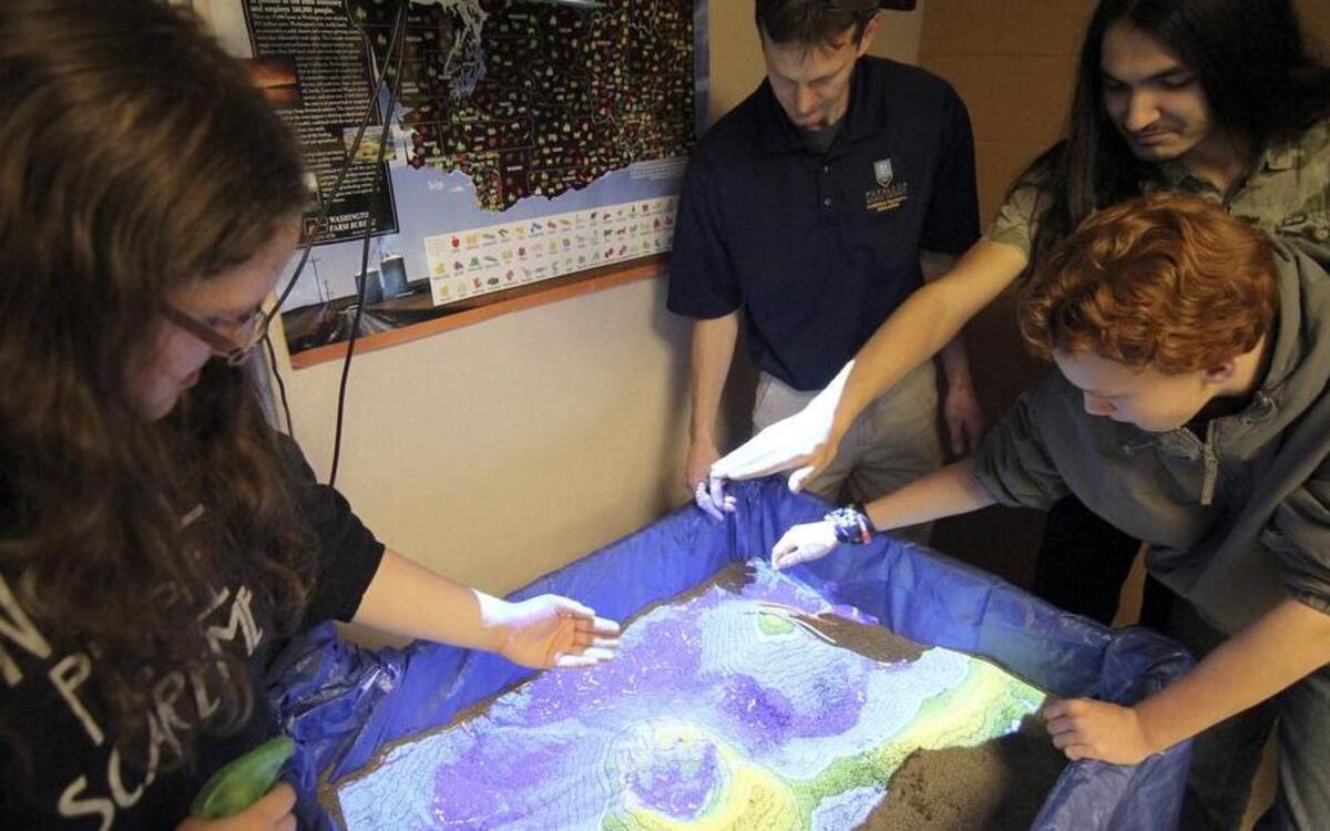 Emerald Ridge High School student Brittany Couts, left, computer science teacher Jason Westerner, and students Josh Arthur and Jacob Waddington work with a computer program that animates topography in real time using projected light to sand. As the students move their hands to create hills, streams, glaciers and other features, the program creates accurate topography modeling through algorithms.