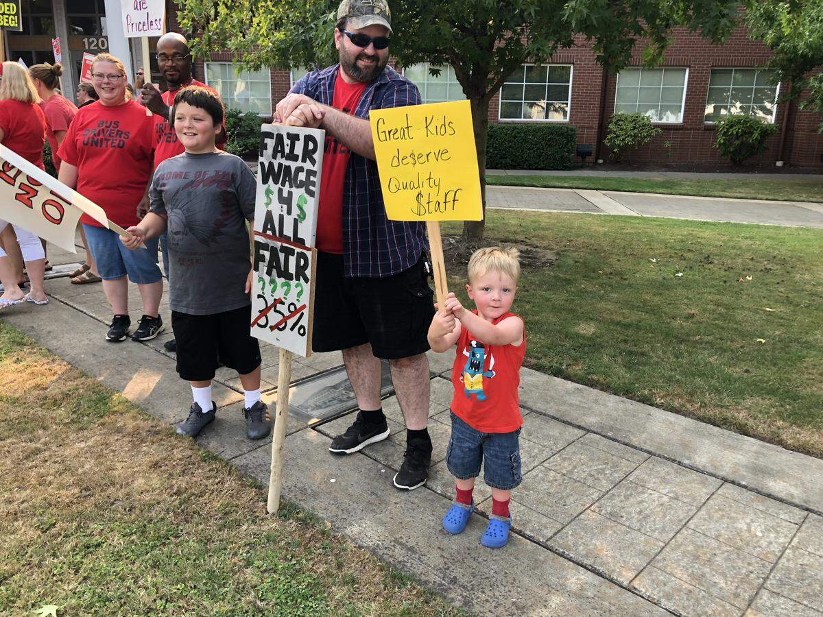 A boy holds a sign that states “Great Kids de$eve Quality $taff” at the Sumner School District rally in support of his father, who wears a “Bus Drivers United” shirt.