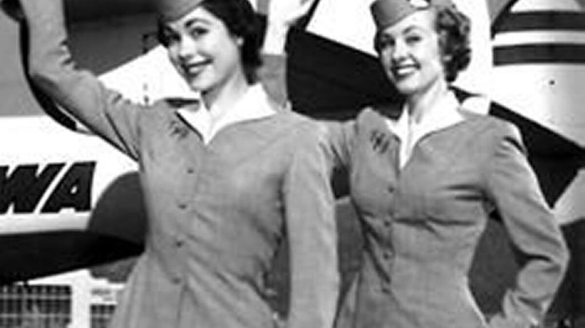 Long-ago TWA flight attendants give their most cheerful five-spot salutes. Those were the good old days, before overbooked seats and passengers being physically “reaccommodated” against their will.
