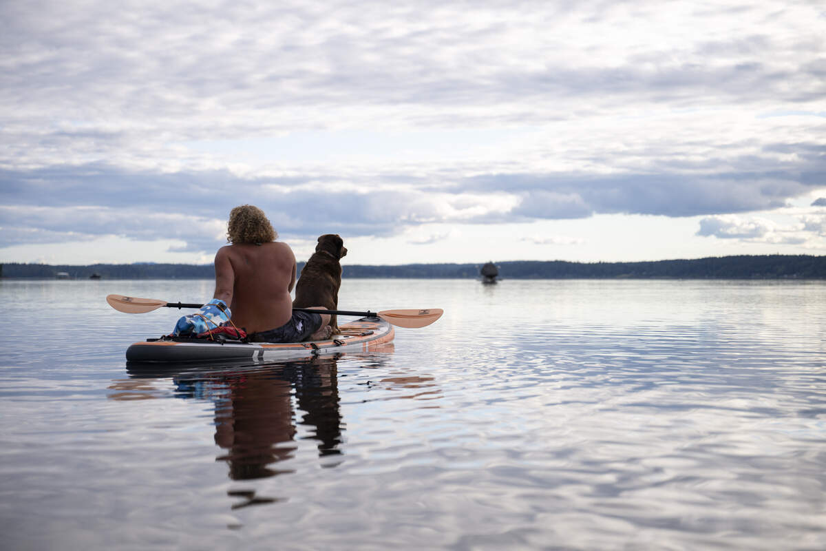 Business owner and outdoor influencer Scott Goerig sits on his paddle board with his dog Rikki-Tikki-Tavi, on Wednesday, June 10, 2026, in Gig Harbor. Goerig, who operates Deadman’s Kayak + SUP Rental & Tours, regularly leads paddlers on day trips out to the sandy shores of Cutts Island State Park, a historic local landmark.